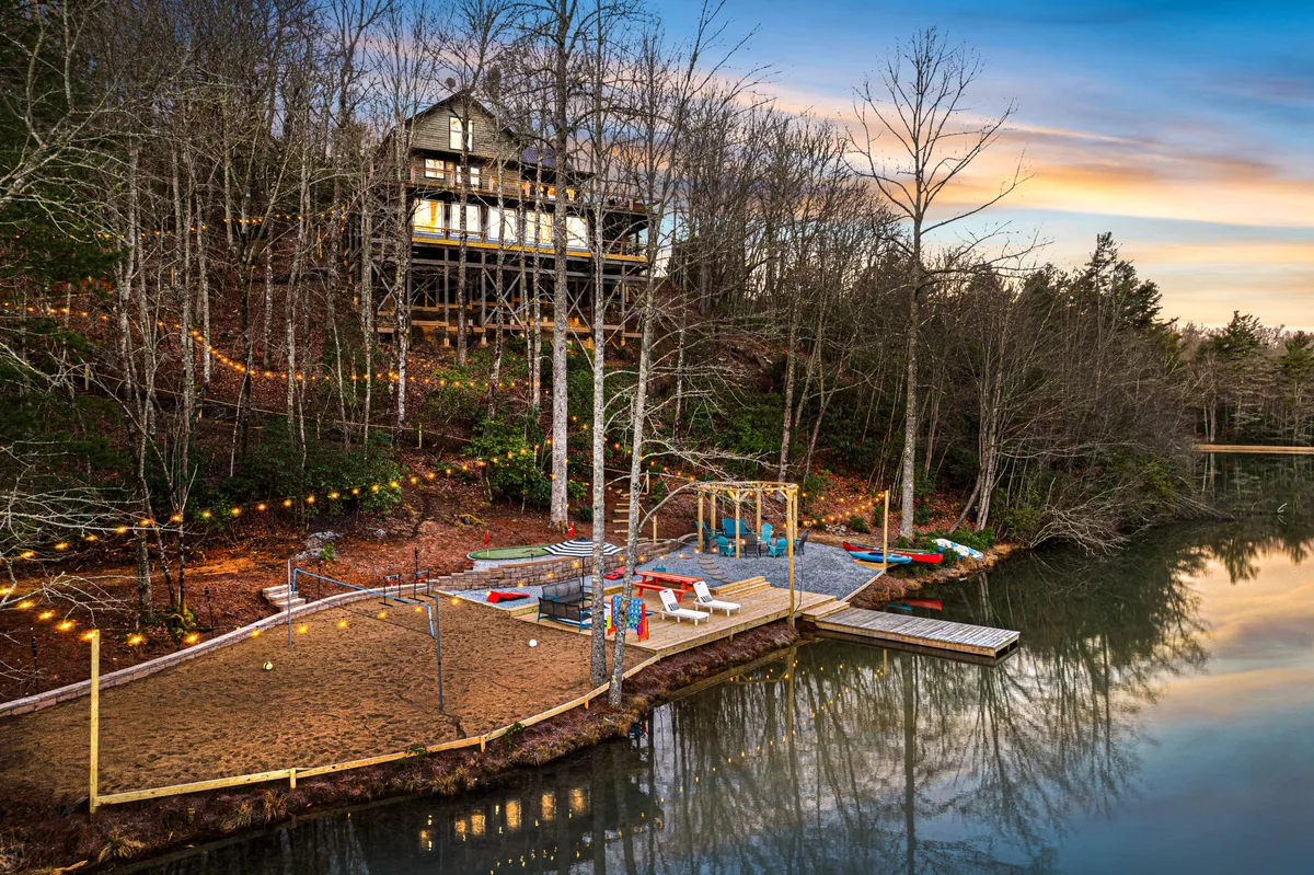 This exterior image captures an elevated house overlooking a sandy beach area, a children's playground, and the serene water surface, all illuminated by gentle daylight. The scene exudes a relaxed, family-friendly atmosphere, with spaces for swimming, fishing, sunbathing, and playground activities visible.