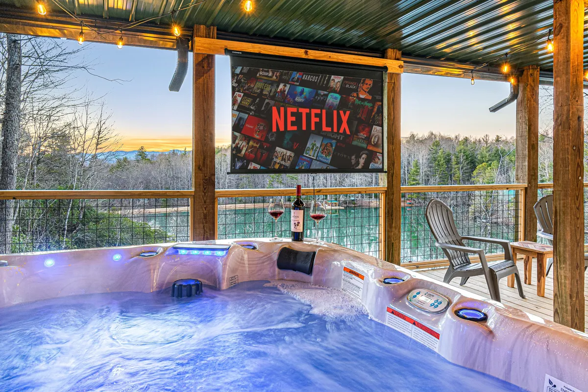 An inviting outdoor hot tub set on a deck, positioned to take in a sweeping landscape, complete with wine glasses and a Netflix screen for ultimate relaxation. The scene feels cozy, luxurious, and ideal for unwinding after a day of wellness activities.