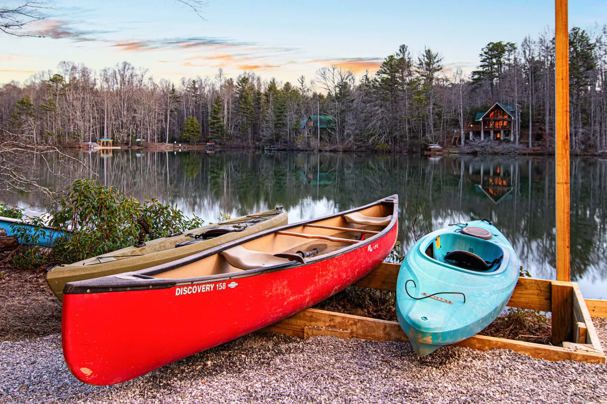 A tranquil lakeside scene featuring a red canoe and blue kayak resting on calm water, surrounded by lush green trees. The lake’s surface is glassy and reflective, suggesting early morning or sunset. The natural shoreline and peaceful setting invite boating, fishing, and quiet contemplation.