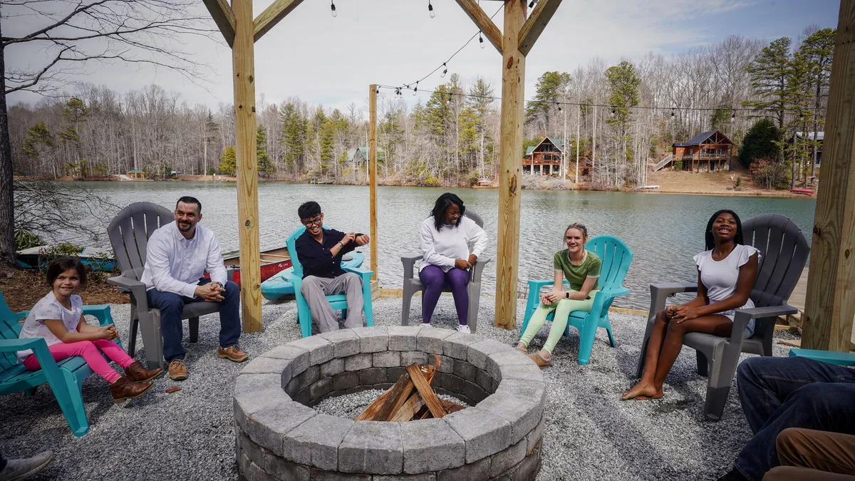 This exterior image shows a group of people gathered around a fire pit next to a lake, with a wooden structure nearby. The sun is setting, casting a warm glow on the water, while everyone enjoys a communal, relaxed atmosphere. Adirondack chairs and outdoor seating enhance the sense of togetherness and comfort.