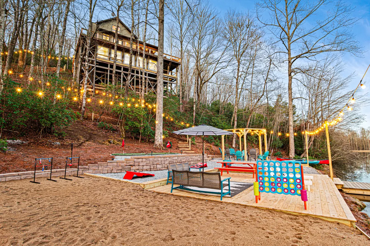 A lively outdoor space with an elevated house, a sandy beach area, colorful outdoor seating, game board, and string lights. The area is perfect for pets to play and families to gather for outdoor fun and relaxation by the water.