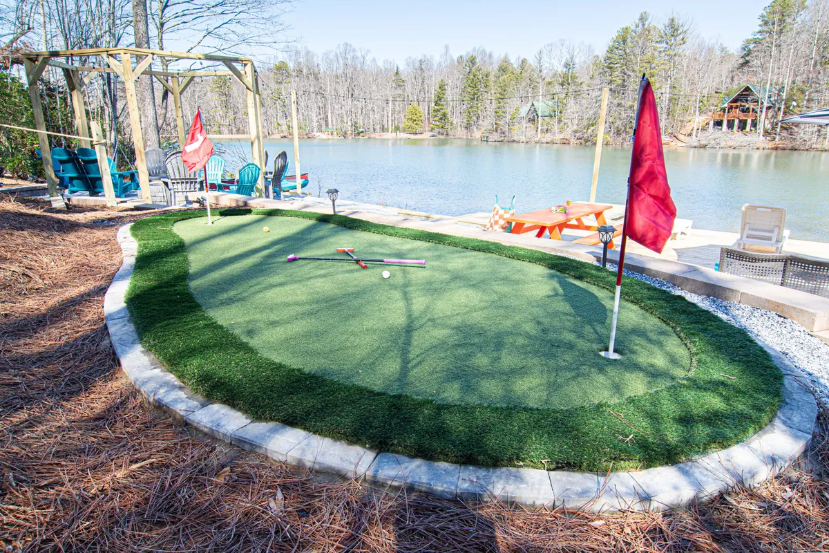 A playful lakeside exterior with a lush putting green, outdoor seating area, flags, golf clubs, and tranquil lake views. The setup is ideal for friendly mini-golf tournaments and socializing with friends while enjoying the scenic water backdrop.