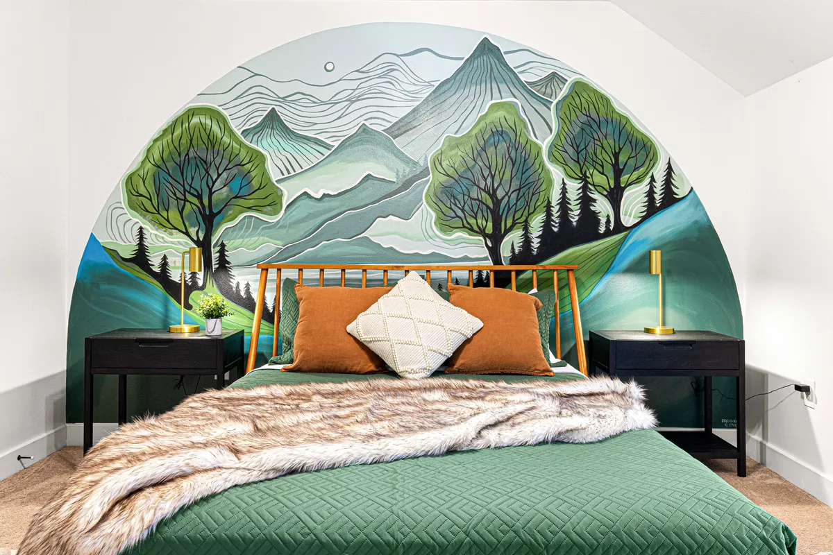 A bedroom with a mural of mountains and trees behind a bed covered in a green quilt and decorative pillows. Nightstands with lamps on either side create an inviting, nature-inspired sanctuary.