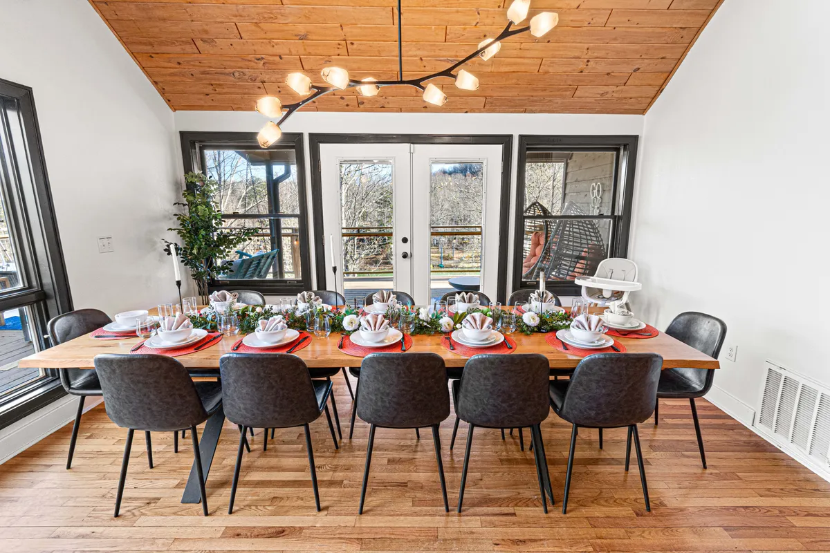 An elegant dining area, highlighted by a large dining table under a modern chandelier, is surrounded by expansive windows bringing in natural light. Decorative plants add warmth and vibrancy, making the space perfect for group meals or celebrations.