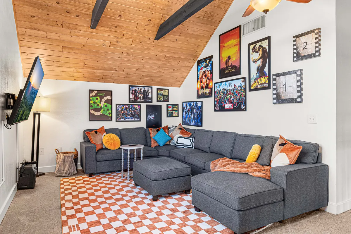 A spacious game lounge or living area with a large sectional sofa, colorful wall art, a plaid rug, and a television. The casual setting is designed for relaxation, socializing, and entertainment, ideal for game nights or movie marathons.