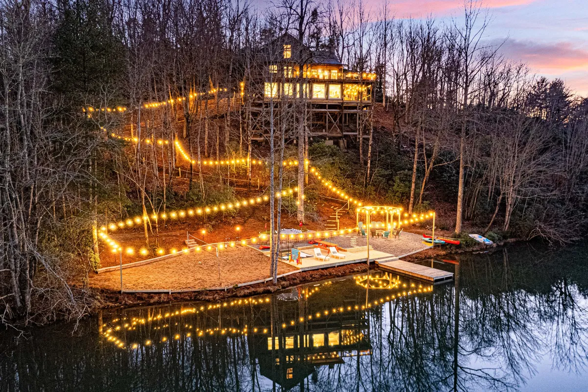 An inviting waterfront scene with an illuminated pathway, a spacious deck above the lake, twinkling lights, and the house perched on a gentle hill. The image conveys a mood of joy and serenity, suggesting group gatherings, outdoor dining, and relaxing by the water in the evening.