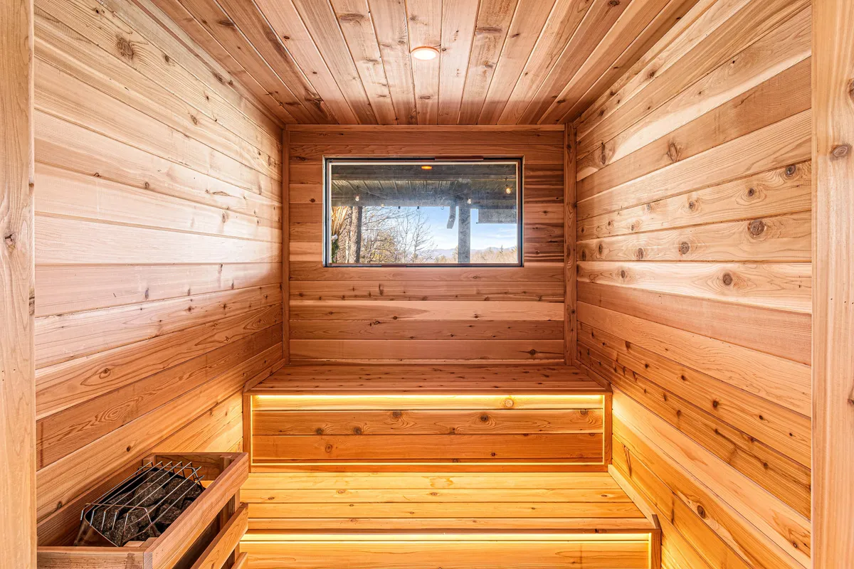 A cozy cedar sauna interior with wooden benches and a window framing the natural landscape, softly illuminated by ambient lighting. The setup invites moments of serene reflection, post-sauna cool-downs, or mindful sessions.