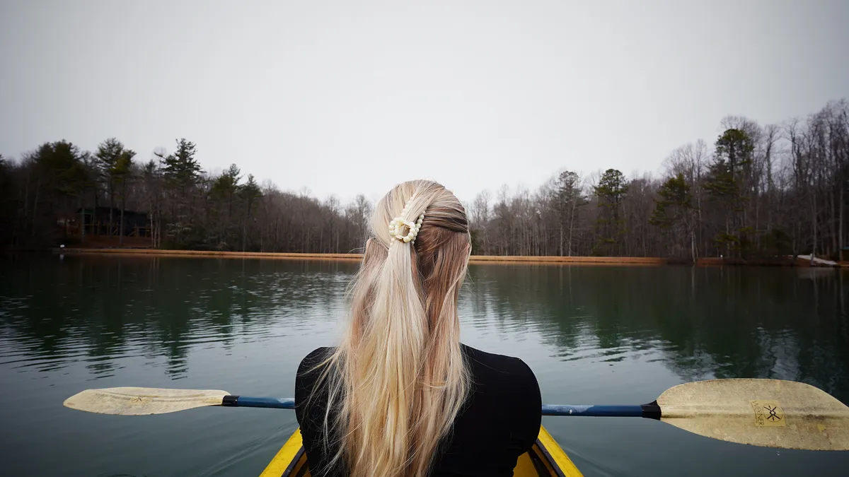 A person kayaks across calm, reflective water bordered by distant trees. The serene lake setting captures the tranquility and adventure of exploring the outdoors, with the gentle ripples and unspoiled nature evoking a sense of peaceful escape.