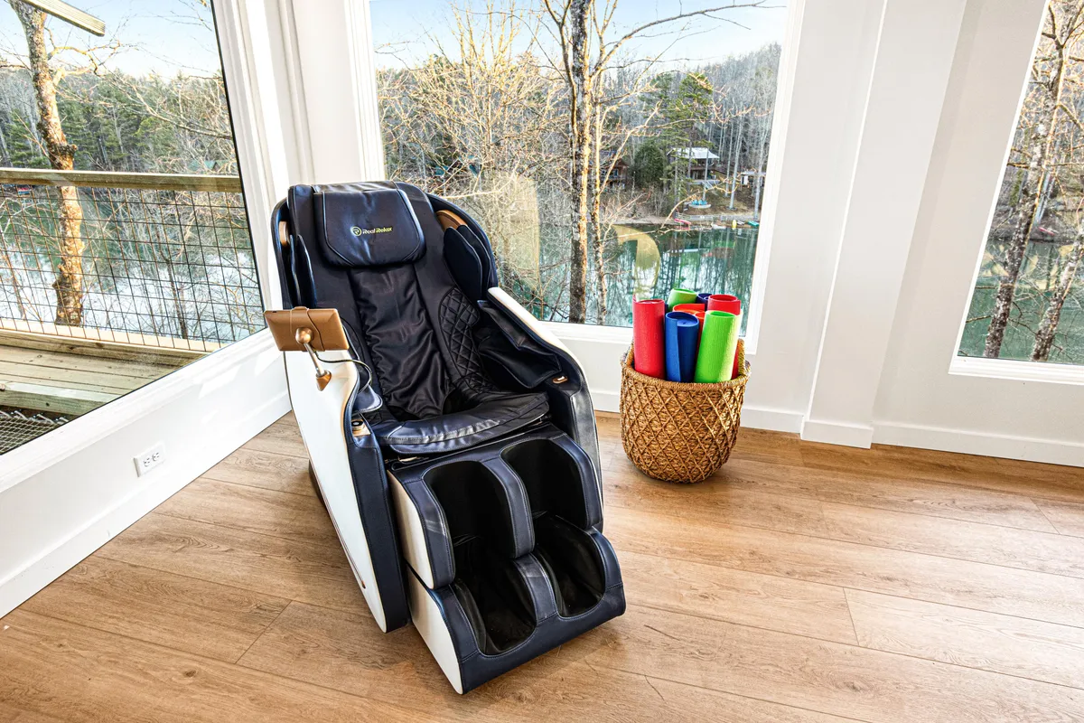 A calm, sunlit interior wellness nook featuring a massage chair with a sweeping view through large windows, accompanied by colorful yoga mats neatly laid out for practice. The space feels soothing and purpose-designed for yoga sessions, meditation, or personal relaxation.