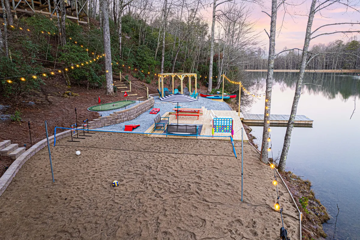 A dynamic lakeside game area featuring a sand volleyball court, kayaks ready by the shore, a colorful play area for kids, decorative lighting, and relaxed lakeside seating. The scene is festive and inclusive, inviting guests of all ages to participate.