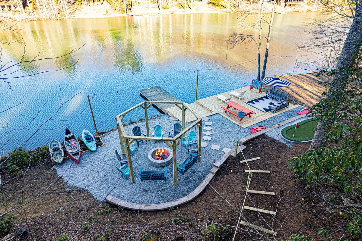 An outdoor scene featuring a fire pit surrounded by Adirondack chairs, colorful kayaks ready by the shoreline, a spacious lawn area, and an outdoor dining setup overlooking the lake. The setting is vibrant and inviting, ideal for both relaxation and active play.