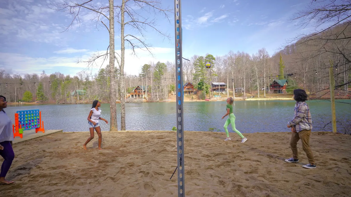 A lively exterior shot of a volleyball net set up by the lake, with people actively playing volleyball and cabins nestled among trees in the background. The vibrant scene exudes energy and togetherness, surrounded by nature.