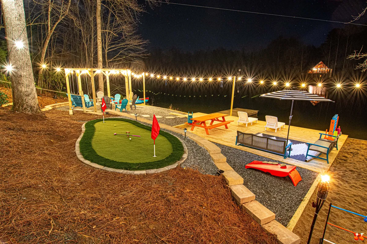 A vibrant exterior shot of a mini-golf green under string lights, surrounded by colorful seating and a sparkling water feature. The setting suggests casual evening fun, friendly competition, and relaxed socializing by the water.