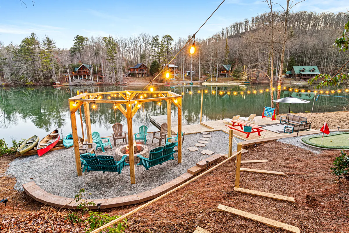 An inviting lakeside setting featuring a gazebo, fire pit, seating arrangements, and a serene view of the water surrounded by trees. The area is designed for communal gatherings, with easy access to the lake for kayaking or fishing adventures.